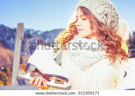 Beautiful caucasian woman going to ice skating outdoor. She dressed in white winter pullover and warm hat. Holding skates shoes. Healthy lifestyle and sport concept at olympic stadium at nature.