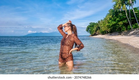 A Beautiful Caucasian Woman With Her Face In Profile, Wearing An Animal Print Swim Cover Up And Long Brown Hair In A Ponytail, Kneeling In Clear Shallow Water Off A Beach On A Tropical Island.