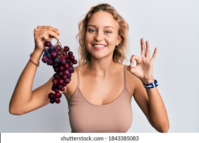 Beautiful caucasian woman holding branch of fresh grapes doing ok sign with fingers, smiling friendly gesturing excellent symbol  - Powered by Shutterstock