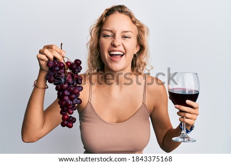 Beautiful caucasian woman holding branch of fresh grapes and red wine smiling and laughing hard out loud because funny crazy joke. 