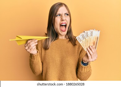 Beautiful Caucasian Woman Holding Paper Airplane And 500 Norwegian Krone Angry And Mad Screaming Frustrated And Furious, Shouting With Anger. Rage And Aggressive Concept. 