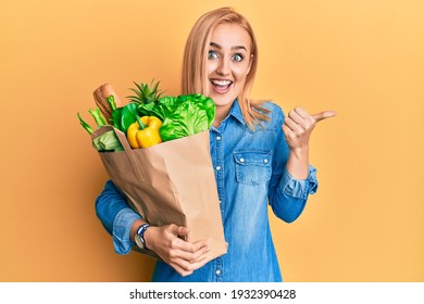 Beautiful caucasian woman holding paper bag with bread and groceries pointing thumb up to the side smiling happy with open mouth 