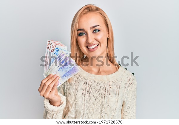 Beautiful caucasian woman holding swedish krona\
banknotes looking positive and happy standing and smiling with a\
confident smile showing teeth\
