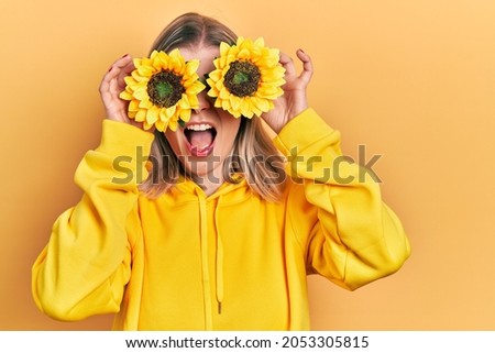 Beautiful caucasian woman holding yellow sunflowers over eyes angry and mad screaming frustrated and furious, shouting with anger. rage and aggressive concept. 