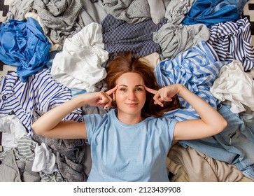 Beautiful Caucasian Woman Smiling And Lying Down With Clutter Clothes On The Floor. Decluttering Pile Of Messy Wardrobe. Cluttered Housewife. Fabric. Shopping. Housework Idea Concept.