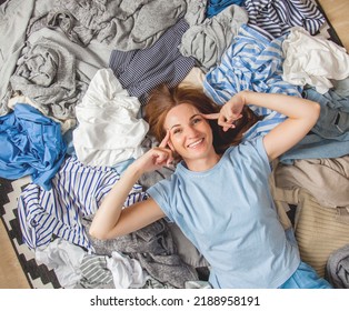 Beautiful Caucasian Woman Smiling And Lying Down With Clutter Clothes On The Floor. Decluttering Pile Of Messy Wardrobe. Cluttered Housewife. Fabric. Shopping. Housework Idea Concept.