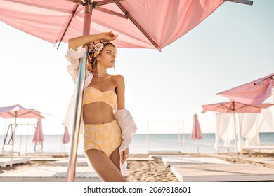 Eine schöne Kaukasierin steht am Strand, voll mit Liegestühlen mit Sonnenschirmen. Herrliche Blondine mit Bandana auf ihrem Kopf in gelbem Badeanzug und weißes Hemd sieht zur Seite.