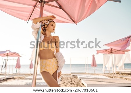 Beautiful caucasian woman is standing on beach crowded with sun loungers with umbrellas. Gorgeous blonde with bandana on her head in yellow swimsuit and white shirt looks to side.