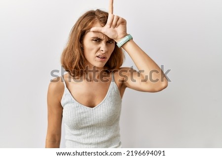 Beautiful caucasian woman standing over isolated background making fun of people with fingers on forehead doing loser gesture mocking and insulting. 