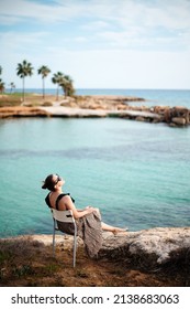 Beautiful Caucasian Woman In Sunglasses And Summer Dress Sitting In Chair On Sea Beach With Palms. Young Happy Brunette Enjoying Vacation On Cyprus