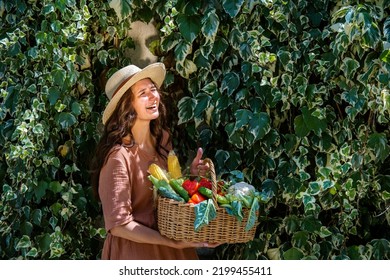 Beautiful Caucasian Woman Wearing Straw Hat And Linen Dress In Garden With Basket Full Of  Vegetables Crop On Ivy Covered Garden Wall.