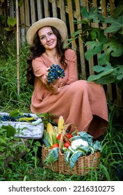 Beautiful Caucasian Woman Wearing Straw Hat And Linen Dress Sitting In Garden With Fresh Grape Berries And Vegetables. Autumn Harvest And Vegetarian Organic Diet.