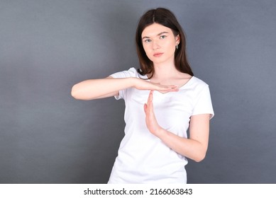 beautiful Caucasian woman wearing white T-shirt over studio grey wall feels tired and bored, making a timeout gesture, needs to stop because of work stress, time concept.