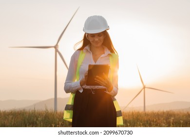 Beautiful Caucasian Woman In White Helmet Working With Digital Tablet At Renewable Energy Farm. Female Inspector Controlling Functioning Of Wind Turbines Outdoors.