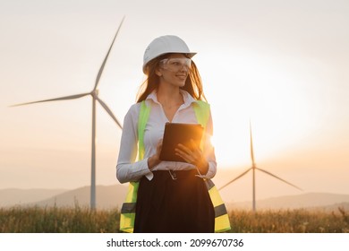 Beautiful caucasian woman in white helmet working with digital tablet at renewable energy farm. Female inspector controlling functioning of wind turbines outdoors.