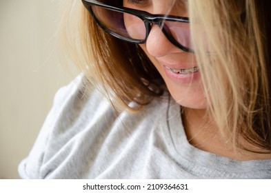 Beautiful Caucasian Young Girl In Braces Looks Down At Phone And Smiles. Brunette Teen Girl With Glasses. Health And Vision Care Concept With Copy Space.