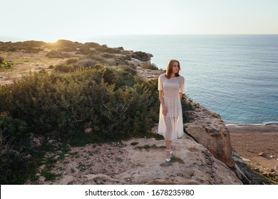 A beautiful caucasian young girl in pink dress stay at the edge of cliff and look to the sunset over the sea - Shutterstock ID 1678235980