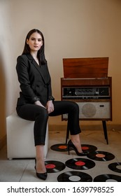 A Beautiful Caucasian Young Woman In A Black Pantsuit Poses Next To A Vintage Record Player.