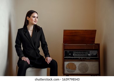 A Beautiful Caucasian Young Woman In A Black Pantsuit Poses Next To A Vintage Record Player.