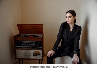 A Beautiful Caucasian Young Woman In A Black Pantsuit Poses Next To A Vintage Record Player.