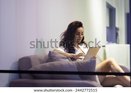 Beautiful Caucasian young woman with long dark hair sitting on sofa in modern living room typing on tablet browsing online. Happy female in white robe using gadget at home Stock fotó © 