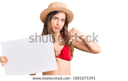 Beautiful caucasian young woman wearing bikini holding blank empty banner with angry face, negative sign showing dislike with thumbs down, rejection concept 