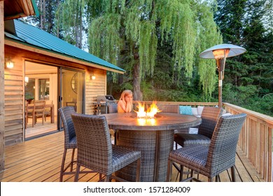 Beautiful Chalet Cider Home Exterior During Evening With Little Girl Sitting Near Fire Pit On  Outdoor Furniture.