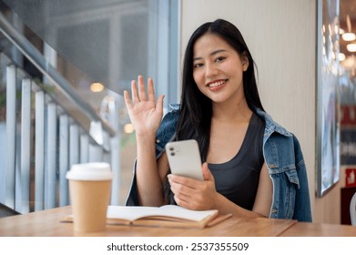 A beautiful, charming Asian woman in denim jacket sitting in a coffee shop, holding a smartphone and smiling and waving her hand at the camera. people and lifestyle concepts