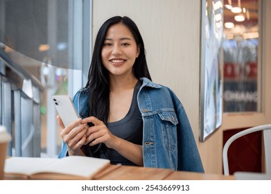 A beautiful, charming Asian woman in denim jacket sitting in a coffee shop, holding a smartphone and smiling at the camera. people and lifestyle concepts - Powered by Shutterstock