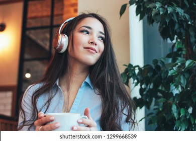 Beautiful Charming Brunette Smiling Asian Girl Enjoys Music In Headphones With Cup Of Coffee Or Tea In Cafe