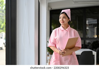 Beautiful Charming Young Asian Female Nurse In A Uniform With Medical Clipboard Stands In The Hospital Hallway. Healthcare Staff Concept