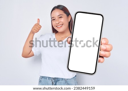 Beautiful cheerful Asian girl wearing casual white t-shirt showing mobile phone with blank black screen and raising thumb up isolated on white background. People lifestyle concept