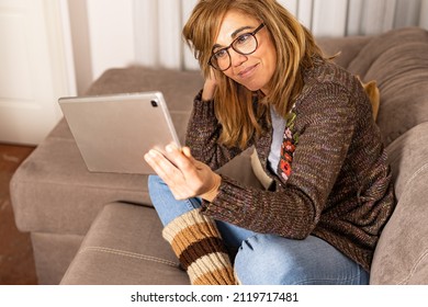 Beautiful Cheerful Blonde Woman Sitting On Sofa At Home Holding Tablet Watching Screen Video Calling Friend Remotely Online Using Video Chat Application.