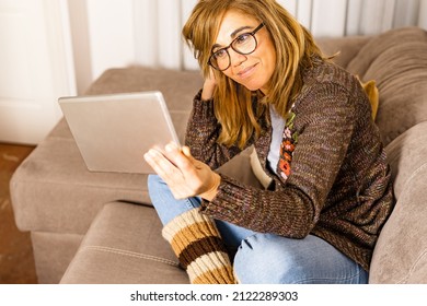 Beautiful Cheerful Blonde Woman Sitting On Sofa At Home Holding Tablet Watching Screen Video Calling Friend Remotely Online Using Video Chat Application.