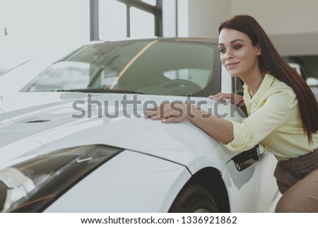 Beautiful cheerful dark haired elegant woman checking out stunning white sportscar at the dealership. Attractive female cusotmer examining car on sale, copy space