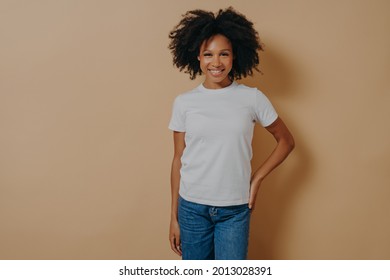 Beautiful Cheerful Dark Skinned Woman In Casual Outfit Smiling At Camera, Cheerful African Female In White Tshirt And Jeans Expressing Positive Emotions, Posing Against Beige Background In Studio