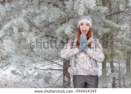 beautiful cheerful happy girl with red hair in a warm hat and scarf playing and fooling around in the snow in the winter forest