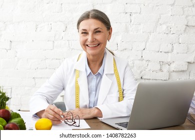 Beautiful Cheerful Mature Sixty Year Old Food Coach Wearing White Overalls And Measure Tape Around Her Neck Smiling Broadly, While Sitting At Desk With Laptop And Vegetables, Having Online Workshop