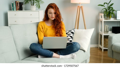 Beautiful cheerful redhead girl using silver laptop while sitting on sofa in living room at home. 
