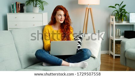 Beautiful cheerful redhead girl using silver laptop while sitting on sofa in living room at home. 