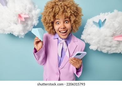 Beautiful Cheerful Woman With Curly Blonde Hair Holds Handmade Paper Aeroplane And Smartphone Dressed In Formal Purple Jacket Works In Office Isolated Over Blue Background White Clouds Overhead.