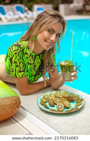 Beautiful cheerful woman smiling in green bikini by swimming pool. Model portrait with cocktail and fruits on summer vacation. Luxury resort
