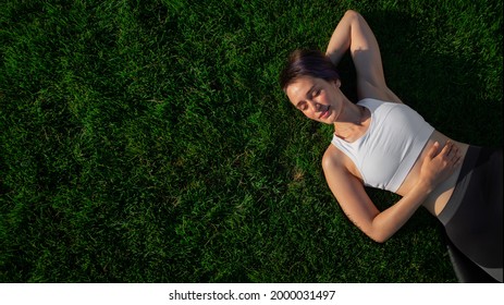 A beautiful, cheerful, cheerful woman in a white T-shirt is lying on the green lawn in the park and smiling. Happy woman relaxing on the grass during sunny summer day. Top view. large format banner