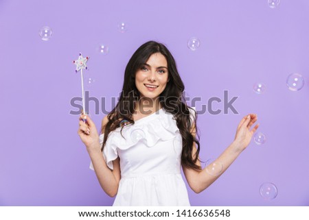 Beautiful cheerful young woman wearing princess outfit standing isolated over violet background, posing with magical wand