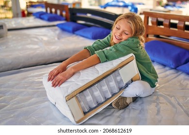 Beautiful child cute girl sitting on the bed and holding a cutaway mattress sample in the store. Purchase of bedding.