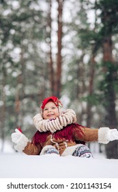 Beautiful child girl has fun with lollipop in hand and bagels bunch on her neck in snowy forest. She dressed in the old Russian style in red headscarf.
