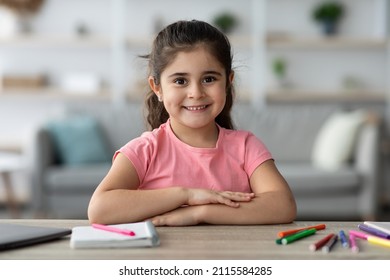 Beautiful Child Portrait. Cute Preteen Arab Girl Posing At Home Interior, Joyful Little Middle Eastern Female Kid Sitting At Desk With Folded Arms, Looking And Smiling At Camera, Copy Space