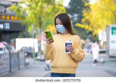 Beautiful Chinese Asian young woman walks on street during sunny autumn day. Holds drinks cup of coffee, takes pictures selfie using mobile cell phone. Wearing face mask to prevent respiratory disease - Powered by Shutterstock