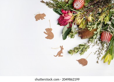 Beautiful Christmas Flat Lay Floral Arrangement Of Mostly Australian Native Flowers, Purple And Red Banksia, Red Waratah And Green Foliage, Decorated By Native Wooden Animals, On A White Background. 