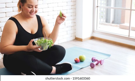 Beautiful Chubby Woman Holding A Bowl Of Salad And Sitting On A Yoga Mat In A Room.